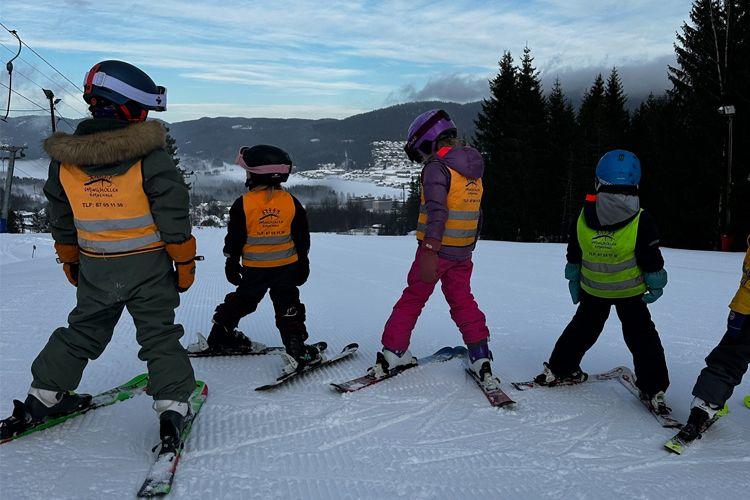 Barn på ski i en bakke, iført fargerike klær og hjelmer, med snødekte trær i bakgrunnen.
