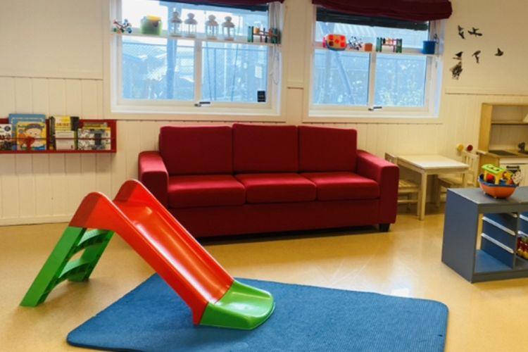Lekeområde med rød sofa, grønn og rød sklie, blått teppe og bokhyller med leker og bøker.