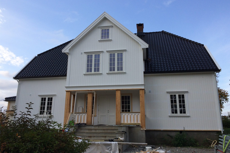 Moderne, hvitt hus med skråtak og tre detaljer, plassert i en hage med busker og byggeutstyr foran.