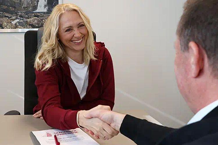 Kvinne med blondt hår smiler mens hun håndhilser på en mann i et kontormiljø.