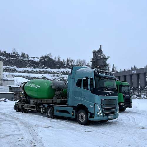 En blå lastebil med betongblander står på snøfylt område ved en steinbrudd. Bakgrunnen viser en annen lastebil og anlegg.