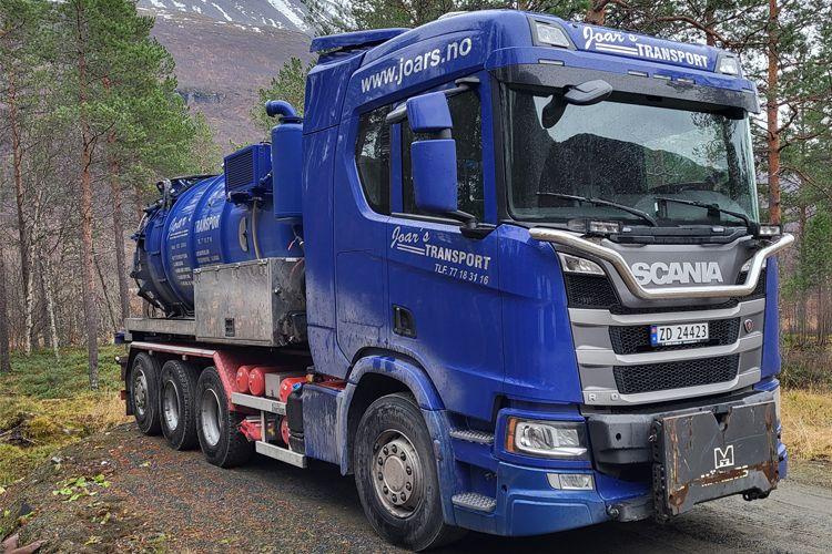 Blå Scania lastebil med tank på tilhenger, parkert i skogsområde med fjell i bakgrunnen.