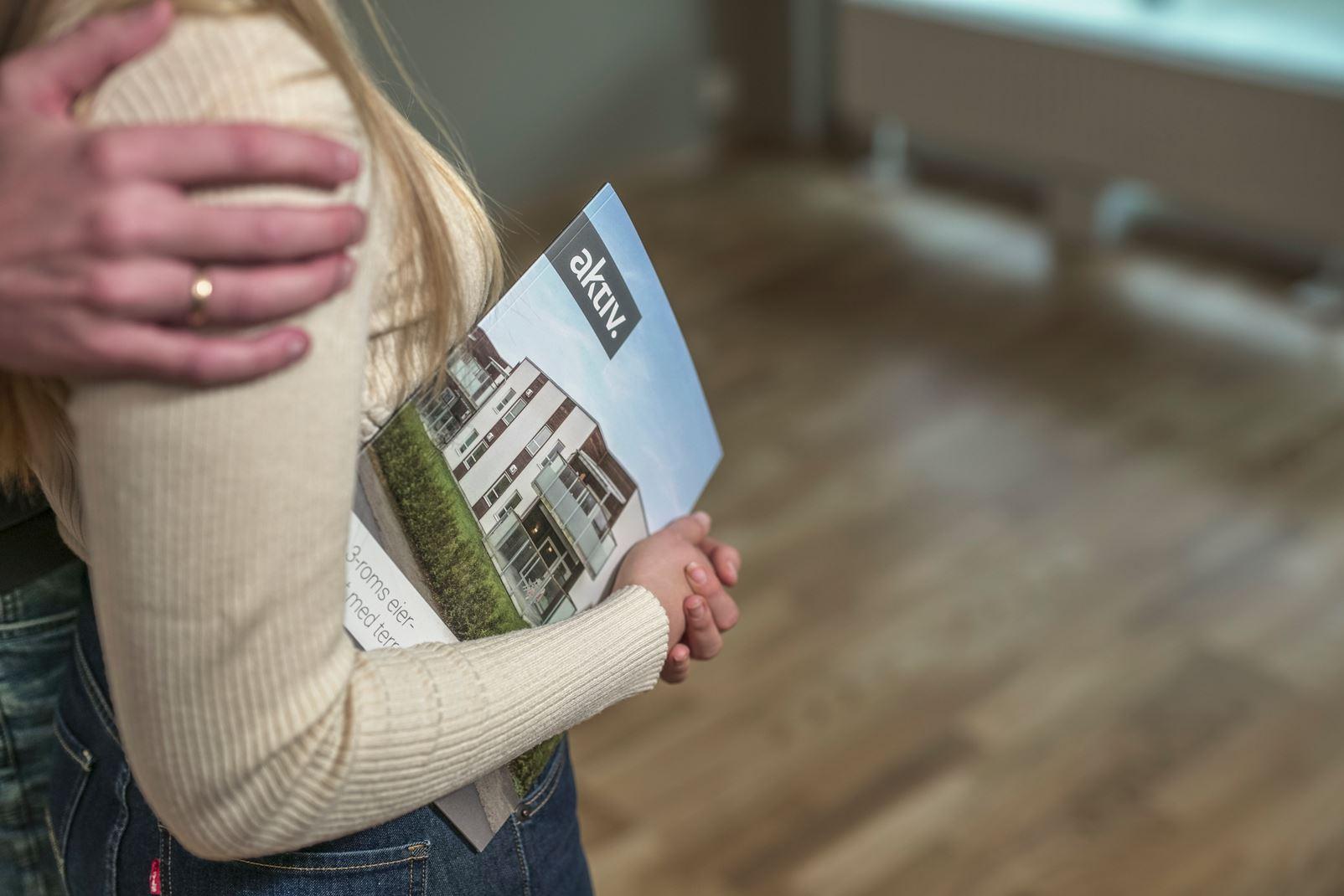 En person holder en boligbrosjyre med et moderne hus på omslaget, mens en annen person står nær dem.