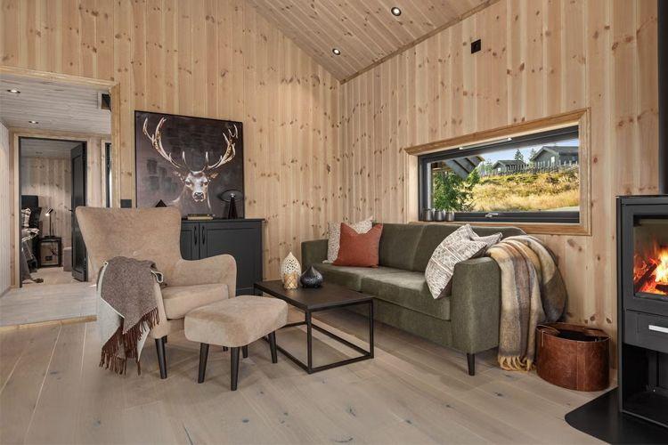Moderne stue med trevegger, grønn sofa, lenestol, peis og kunstverk av en hjort på veggen.
