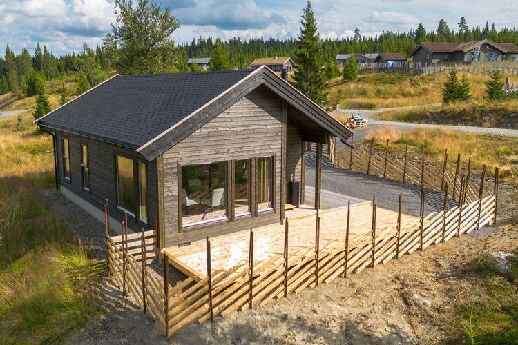 Moderne hytte med stor terrasse og gjerde, omgitt av skog og fjell i bakgrunnen.