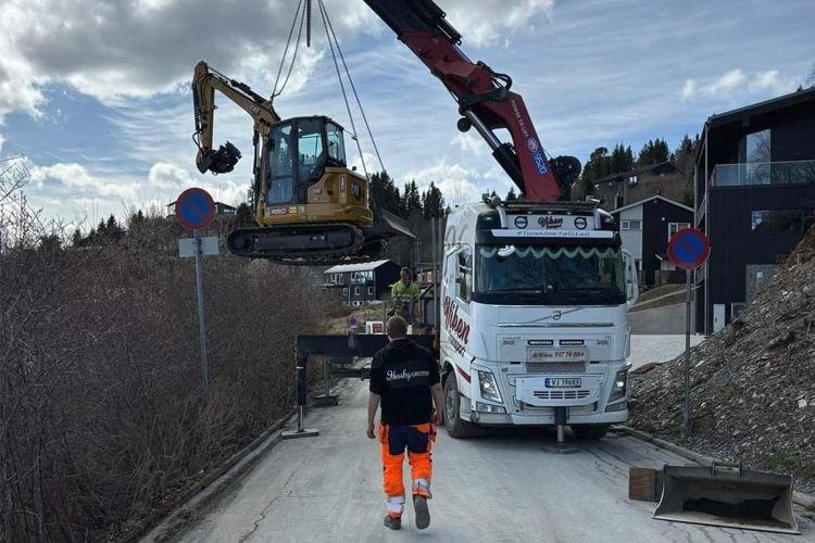 En gravemaskin heises opp av en kran på en vei, med en arbeider i oransje arbeidsklær i forgrunnen.