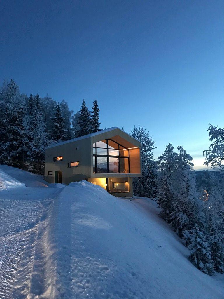 Moderne hytte i snødekt landskap med store vinduer og lysende interiør, omgitt av trær.