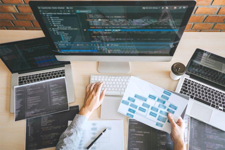 En person som skriver på et tastatur med flere datamaskiner og dokumenter med koding og diagrammer på et skrivebord.