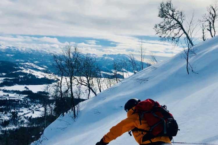 En person i gul jakke klatrer ned en snødekt bakke med fjell og daler i bakgrunnen.