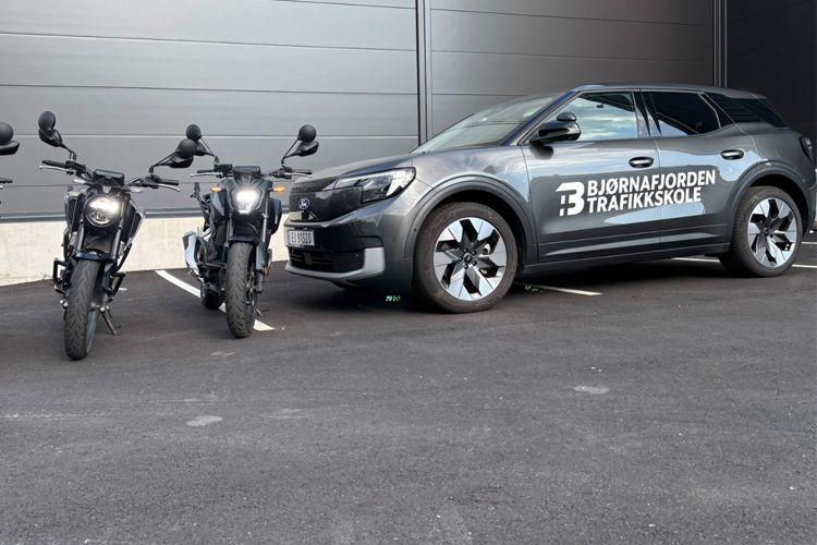 To motorsykler og en bil parkert ved en bygning, med teksten "Bjørnafjorden Trafikkskole" på bilen.