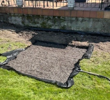 Et område med grus dekket av svart plast, plassert på en gressplen ved en murvegg.