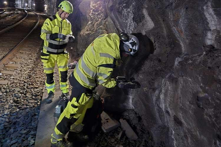 To personer i gule arbeidsklær jobber med en bergvegg ved jernbanelinjen om natten.
