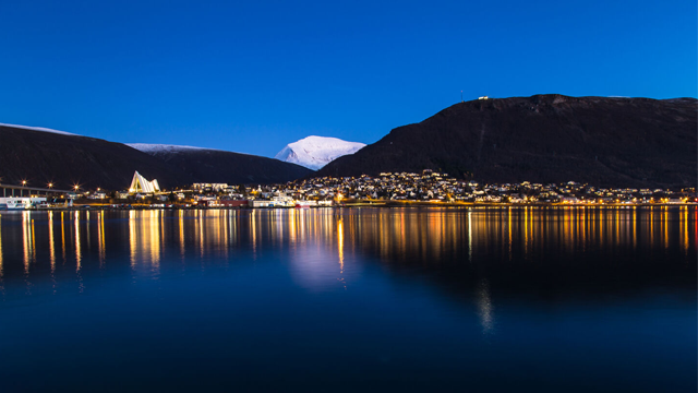 Nattbilde av en by ved fjorden med opplyste bygninger og snødekte fjell i bakgrunnen.