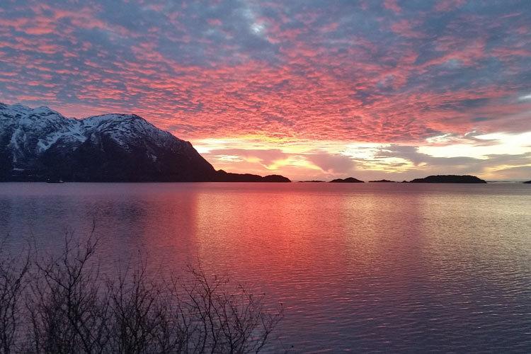 Fargerik solnedgang over fjorden med snødekte fjell i bakgrunnen og rolig vann foran.