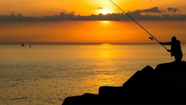 Fisker som står på steiner og fisker ved solnedgang over havet med seilbåter i bakgrunnen.