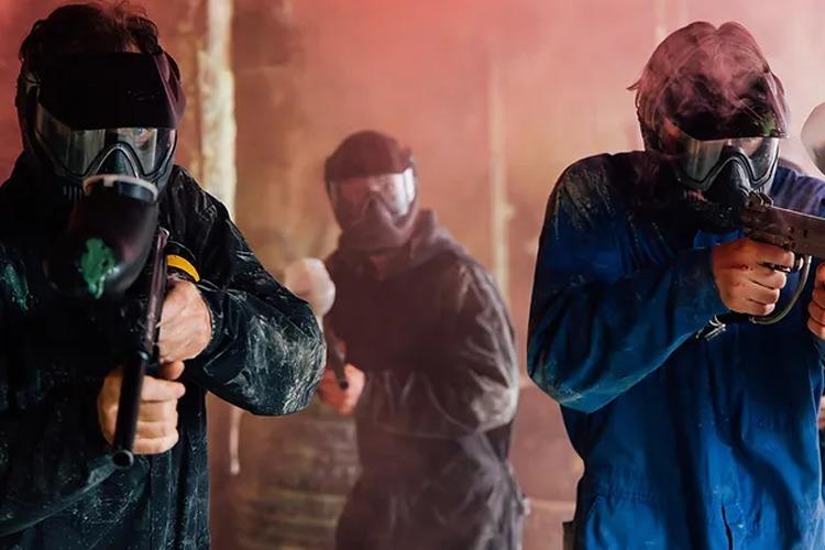 Tre personer i beskyttelsesutstyr spiller paintball i en røykfylt arena.