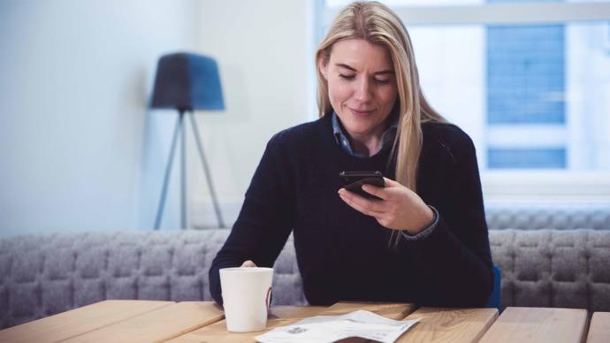 Kvinne sitter ved et bord med en kopp kaffe og ser på mobilen sin i et lyst rom.