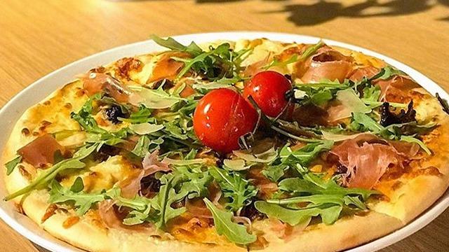 En pizza med ruccola, skinke, oliven og cherrytomater på toppen, servert på en hvit tallerken.