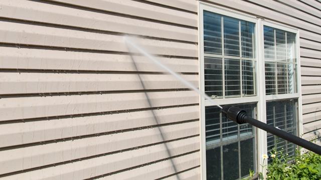 Vannspray fra en høytrykkspyler som rengjør en vegg med vinduer i bakgrunnen.