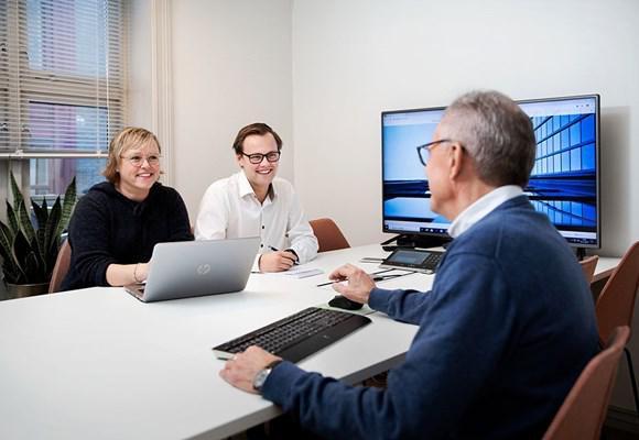 To personer sitter ved et bord med en laptop, mens en annen person snakker til dem i et moderne kontor.