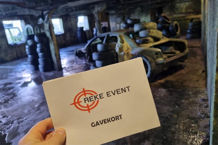 Hånd som holder et gavekort fra Reike Event foran en gammel bil i et rom med dekkstable.