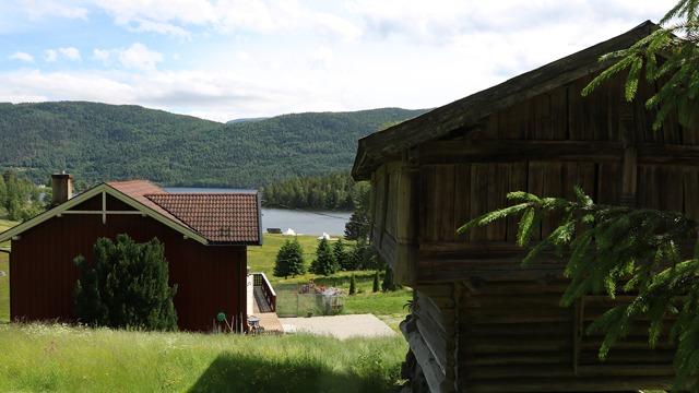 Utsikt over en rød hytte ved en innsjø, omringet av grønne trær og fjell i bakgrunnen.