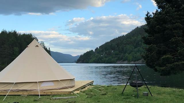 En glamping-telt ved en innsjø, omringet av fjell og trær, med en vakker himmel i bakgrunnen.