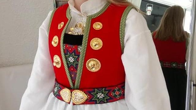 Kvinne iført tradisjonell bunad med rød vest, hvit skjorte og dekorativt belte med gullknapper.