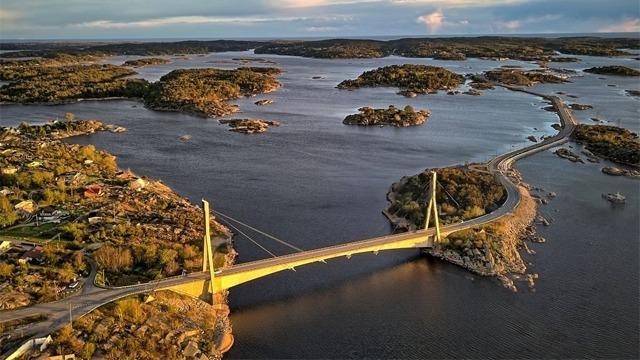 En bro med hengende kabler som krysser en elv, omgitt av øyer og vann i et naturskjønt landskap.