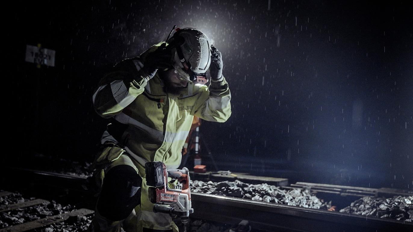 En gruvearbeider i regn, iført hjelm og arbeidsklær, står ved jernbaneskinner i mørket.