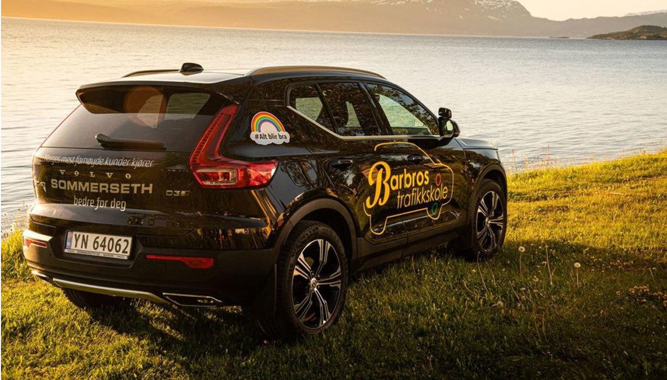 En svart SUV med reklame står på gresset ved sjøen, med fjell i bakgrunnen og solnedgang.