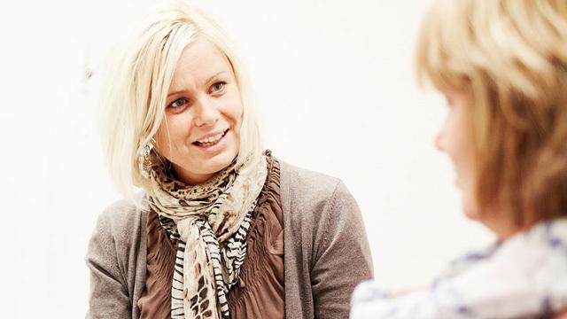 To kvinner i samtale, den ene med blondt hår og skjerf, den andre med brunt hår, begge smilende.