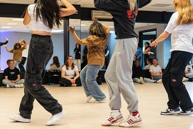 Fire dansere i bevegelse under en danseøkt, med en gruppe tilskuere i bakgrunnen.