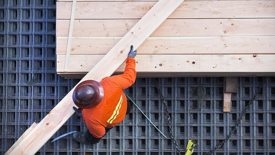 En arbeider i oransje jakke som monterer treplanker på en byggeplass med plastunderlag.