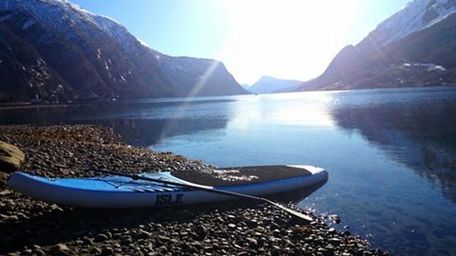 En stand-up paddleboard ligger på en steinete strand ved en fjord, omkranset av fjell i sollys.