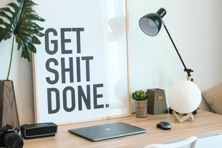 Moderne arbeidsplass med en plakat som sier "GET SHIT DONE", laptop, lampe og planter på skrivebordet.