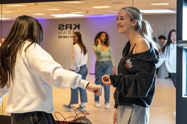 To jenter danser og smiler i et rom med speil og moderne belysning. Flere personer er synlige i bakgrunnen.