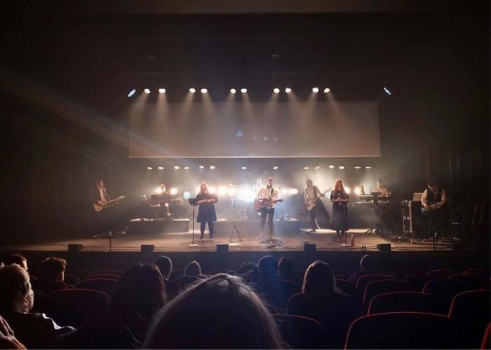 En konsert med musikere på scenen, opplyst av sterke lys, med publikum som ser på fra setene.