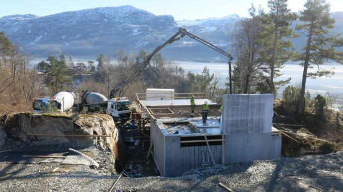 Byggeplass med en betongpumpe som leverer betong til en ny bygning, omgitt av fjell og trær.