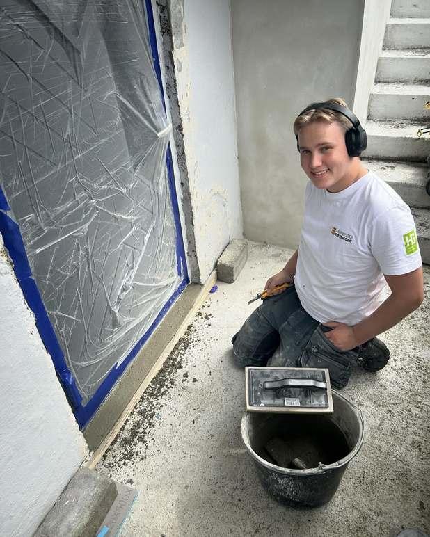 En håndverker jobber med flislegging ved en dør, med verktøy og en bøtte ved siden av seg.