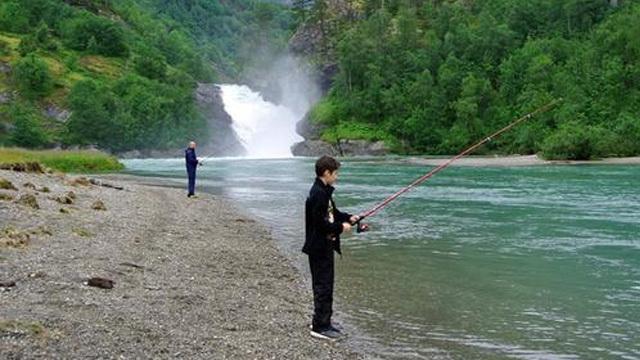 To personer fisker ved en elv med en foss i bakgrunnen, omringet av frodig grønn natur.