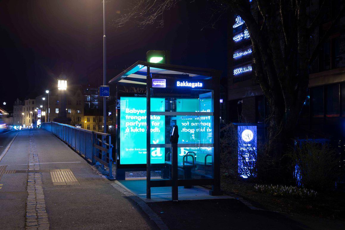 Lysende busstopp med reklame og blå belysning på en nattlig gate.