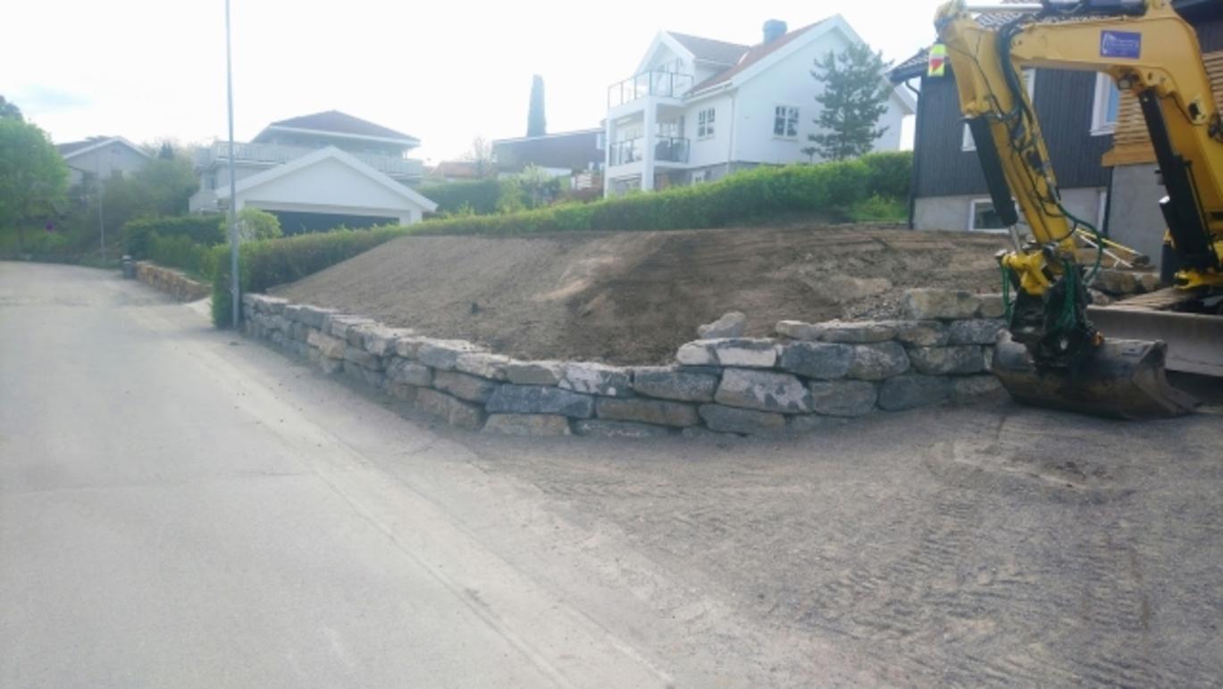 En byggeplass med en gravemaskin og en steinmur langs veien, omgitt av hus og grøntområder.