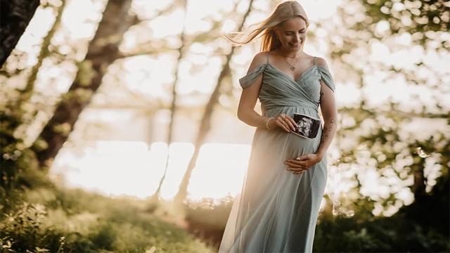 Gravid kvinne i en lys kjole som holder magen sin, står i en solfylt skog med trær i bakgrunnen.
