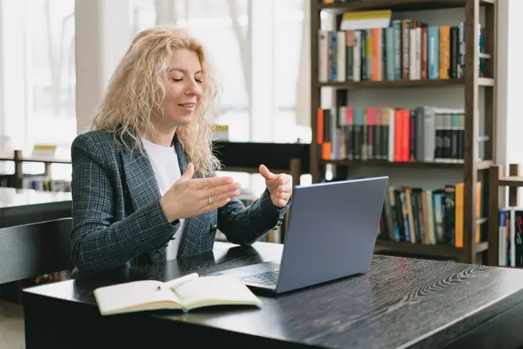 Kvinne med krøllete hår sitter ved et bord i et bibliotek og har videomøte på laptop.