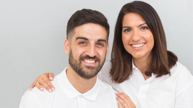Et smilende par i hvite skjorter, poserer sammen foran en lys grå bakgrunn.