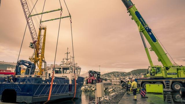 En kran heiser en båt ved havnen, med arbeidere og annet utstyr synlig i bakgrunnen.