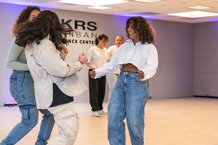 To dansende personer i en lys dansestudio, iført avslappede klær og smilende, med andre dansere i bakgrunnen.