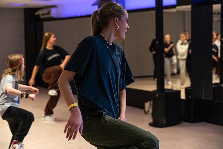 Ung jente danser i en speilsal med andre dansere i bakgrunnen, fokusert på bevegelsene sine.