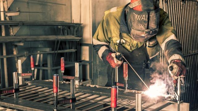 En sveiser i arbeid, iført verneutstyr, sveiser metallplater med gnister som spruter.